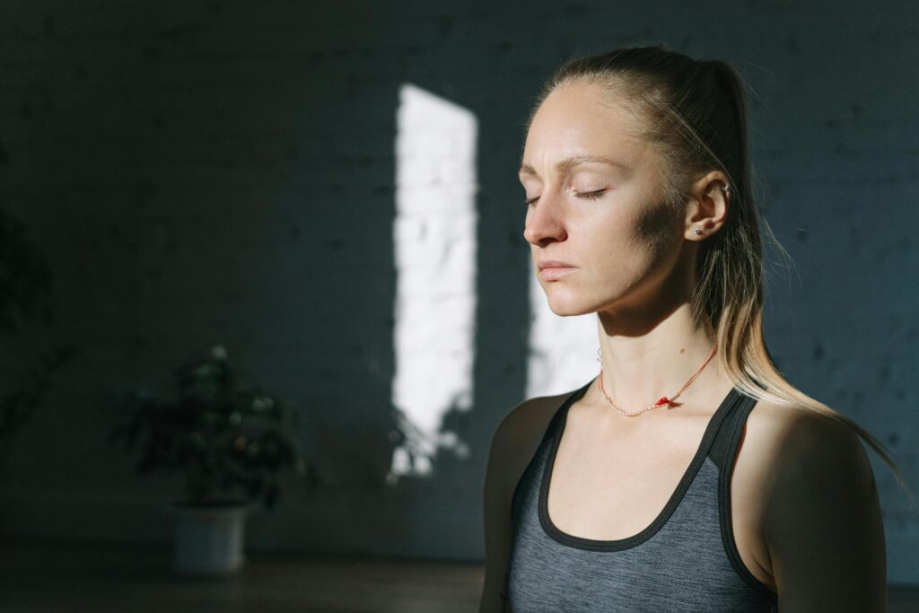 A woman in meditation inside a sunlit room, eyes closed, feeling calm and peaceful.