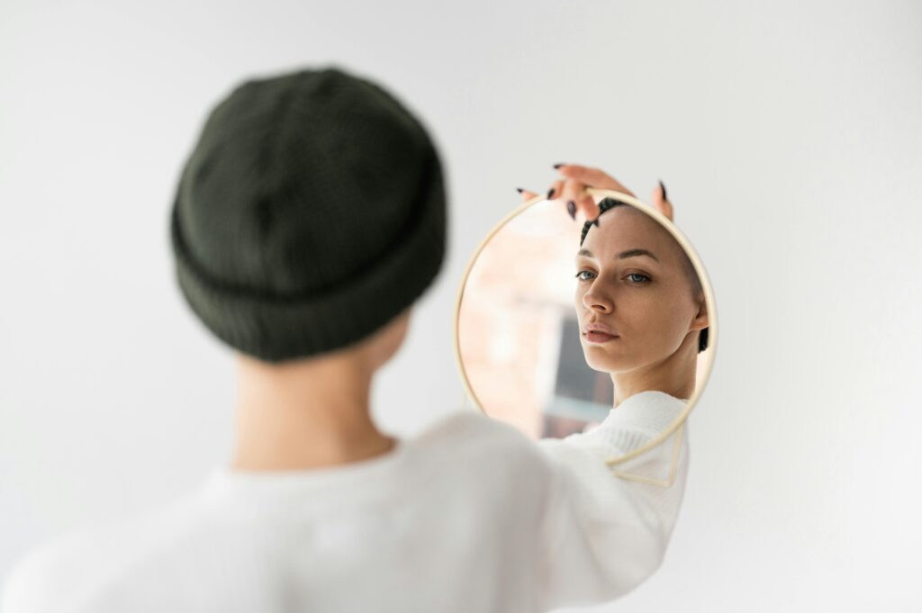 Back view of self assured young female in casual clothes and hat looking in round cosmetic mirror against white background